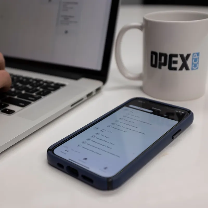 A laptop and a phone open to a client's program, beside an OPEX CCP mug